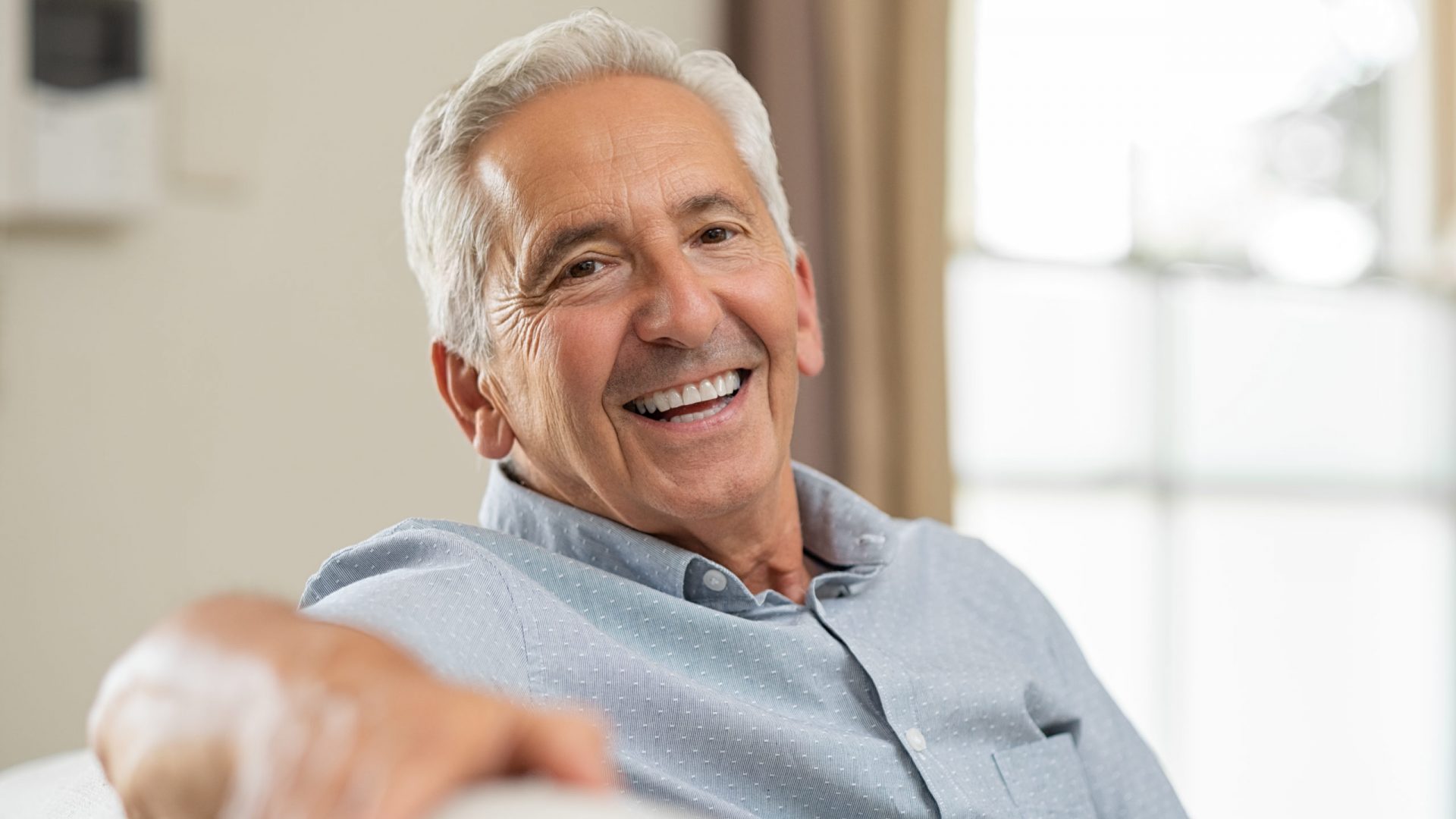 Portrait of happy senior man smiling at home. Old man relaxing on sofa and looking at camera. Portrait of elderly man enjoying retirement.