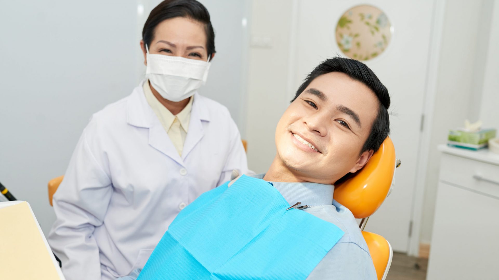 Asian man sitting in dental chair in office with smiling professional doctor in gown and mask near looking at camera